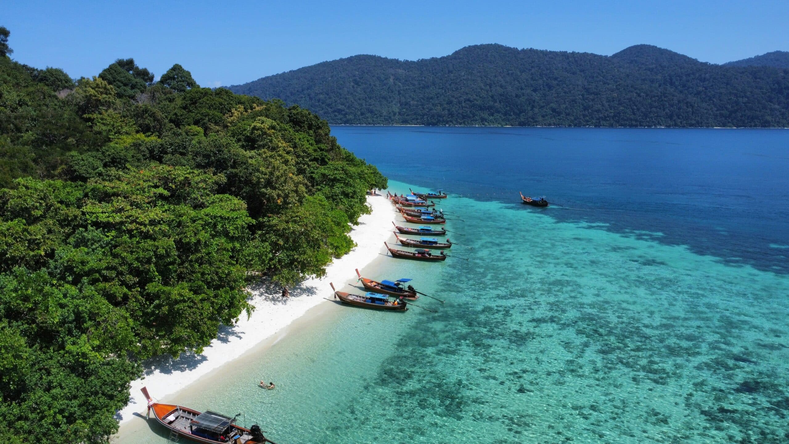Aerial view of a tropical beach on Koh Lipe with lush green trees to the left, white sand, and turquoise water. Traditional long-tail boats line the shore. Hills rise in the distance under a clear blue sky, showcasing why many consider this paradise the best time to visit.