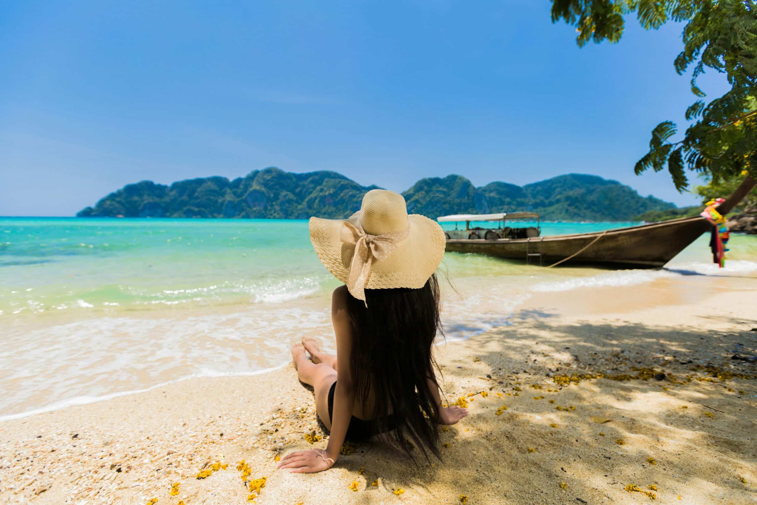 A woman wearing a sun hat sits on a sandy beach in Phuket, facing the turquoise sea. A boat is anchored nearby, and lush green islands are visible under the clear blue sky. The best time to visit Phuket ensures such serene views and perfect weather for relaxation.