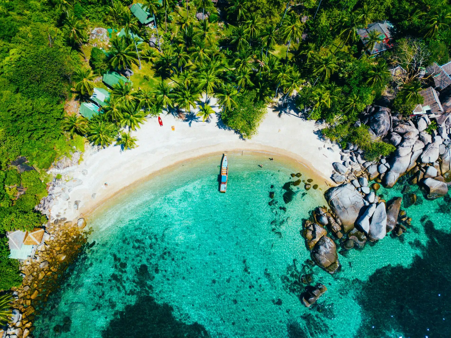 Aerial view of a tropical beach with clear turquoise water. A boat is anchored near the shore, and palm trees line the sandy beach. For those wondering about the best time to visit Koh Tao, rocks are visible in the water alongside several small buildings nestled among the trees.