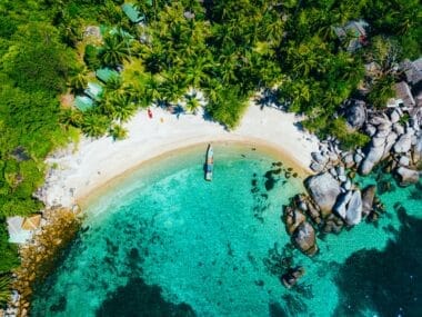 Aerial view of a tropical beach with clear turquoise water. A boat is anchored near the shore, and palm trees line the sandy beach. For those wondering about the best time to visit Koh Tao, rocks are visible in the water alongside several small buildings nestled among the trees.