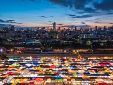 Amid the vibrant night market, where rows of colorful, illuminated stalls beckon visitors, the season unfolds under a darkening blue and orange sky. In this lively place, tall buildings in the background are silhouetted against the breathtaking sunset.