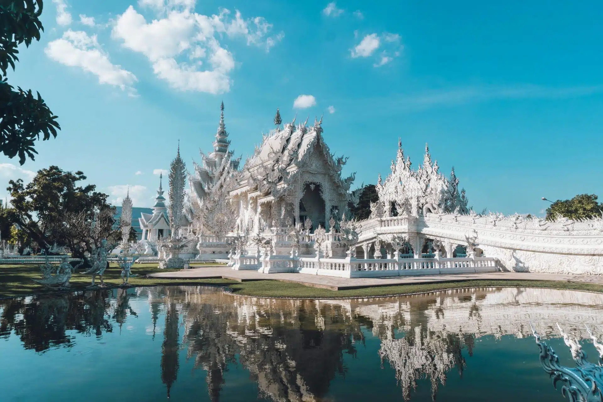 The image captures the White Temple (Wat Rong Khun) in Chiang Rai, Thailand. This ornate white masterpiece, mirrored beautifully in a reflecting pond, displays intricate designs against a clear sky with few clouds. Trees gracefully surround the temple.
