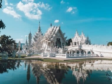 The image captures the White Temple (Wat Rong Khun) in Chiang Rai, Thailand. This ornate white masterpiece, mirrored beautifully in a reflecting pond, displays intricate designs against a clear sky with few clouds. Trees gracefully surround the temple.