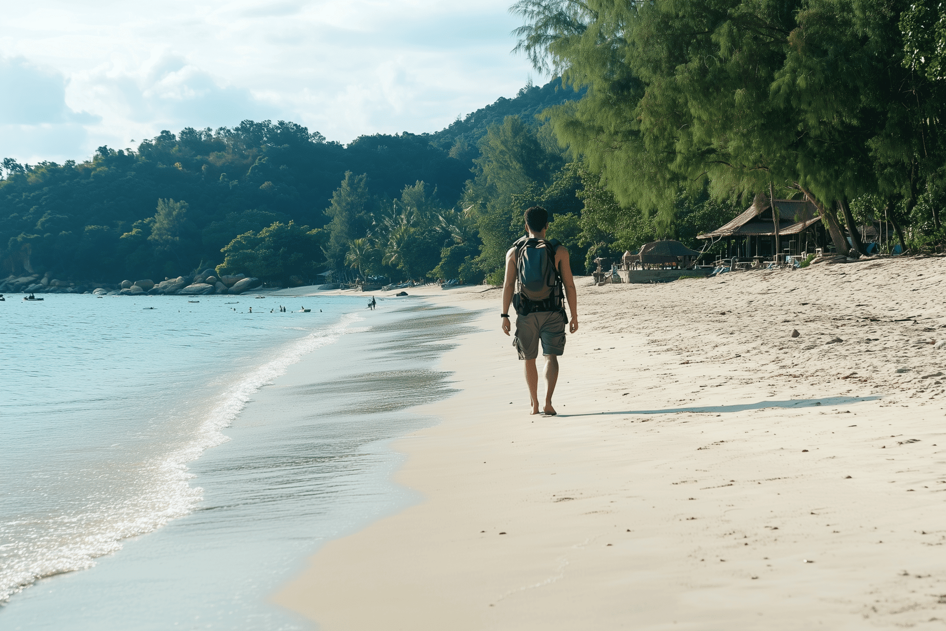 A person with a backpack strolls along a serene beach, gentle waves lapping the shore. Lush green hills and trees frame the scene while rustic wooden huts peek on the right. Its an idyllic view, hinting at why many say the best time to visit Ko Samet is during tranquil moments like these.
