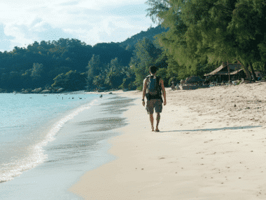 A person with a backpack strolls along a serene beach, gentle waves lapping the shore. Lush green hills and trees frame the scene while rustic wooden huts peek on the right. Its an idyllic view, hinting at why many say the best time to visit Ko Samet is during tranquil moments like these.