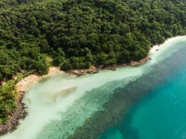 Aerial view of a lush green tropical island with a dense forest and a secluded sandy beach. Clear turquoise water surrounds the island, revealing a coral reef near the shoreline. A few small buildings are visible among the trees, making it the best time to visit Koh Chang before the rain arrives.