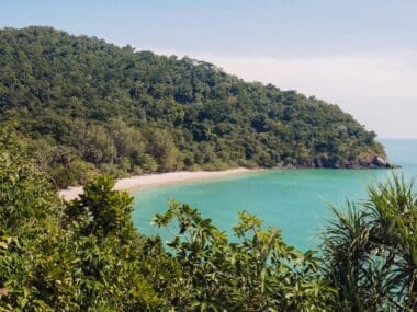 A scenic view of a lush, green hillside on a serene island leads to a sandy beach beside turquoise waters. The hill is covered with dense tropical vegetation under a clear blue sky.