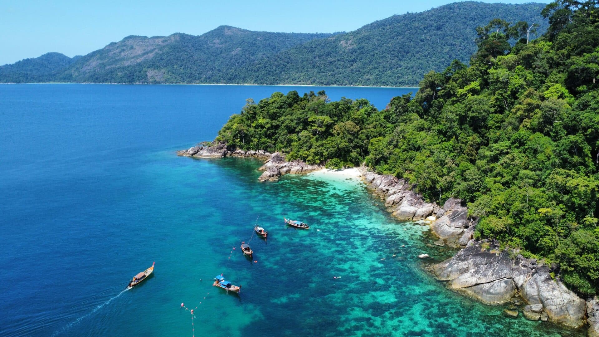 Aerial view of tropical Koh Lipe showcases lush green trees, turquoise and deep blue waters, rocky coastline, and small boats floating nearby. The distant hills are covered with dense forest under a clear blue sky, the best time to visit Koh Lipe.