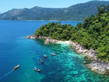 Aerial view of tropical Koh Lipe showcases lush green trees, turquoise and deep blue waters, rocky coastline, and small boats floating nearby. The distant hills are covered with dense forest under a clear blue sky, the best time to visit Koh Lipe.