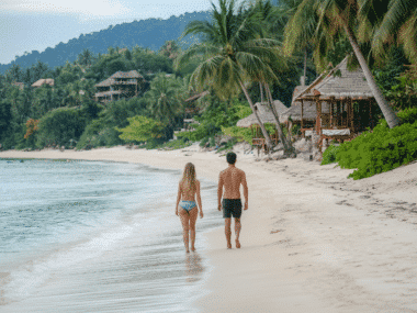 A couple strolls along a tropical beach on Koh Samui, surrounded by clear blue water and lush greenery. Wooden huts line the shoreline as palm trees sway gently under a partly cloudy sky, revealing why many claim this is the best time to visit.