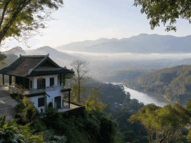 A serene landscape features a house on a hillside in Chiang Rai, overlooking a river winding through the lush valley of the Golden Triangle. Mist drapes over distant wooded mountains under a clear blue sky, creating a peaceful and picturesque scene reminiscent of Wat Rongs elegance.