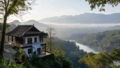 A serene landscape features a house on a hillside in Chiang Rai, overlooking a river winding through the lush valley of the Golden Triangle. Mist drapes over distant wooded mountains under a clear blue sky, creating a peaceful and picturesque scene reminiscent of Wat Rongs elegance.