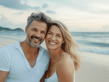 A smiling couple poses for a photo on a beach during their trip. The man has salt-and-pepper hair and a beard, wearing a white shirt, while the woman with long blonde hair wears a white tank top. The ocean and a cloudy sky are in the background.