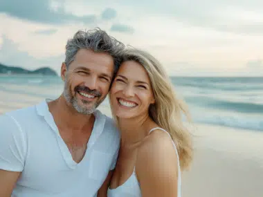 A smiling couple poses for a photo on a beach during their trip. The man has salt-and-pepper hair and a beard, wearing a white shirt, while the woman with long blonde hair wears a white tank top. The ocean and a cloudy sky are in the background.