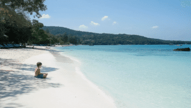 A child sits on a pristine white sandy beach by turquoise waters under a clear blue sky. Beyond, lush green hills of the national park provide a breathtaking view, while a few people stroll along Ko Samets tranquil shoreline.