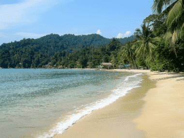 A serene tropical beach on Koh Chang boasts golden sand, gentle waves, and clear blue water. Lush green hills and a few small buildings are in the distance. Palm trees line the shoreline under a bright blue sky, making it the perfect holiday escape.