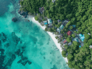 Aerial view of Sairee Beach with turquoise waters and coral reefs. Lush green forest surrounds small, colorful buildings, while palm trees line the white sandy shore. A lone boat is anchored near the beach, adding to the serene beauty of this tropical paradise.