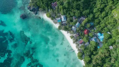 Aerial view of Sairee Beach with turquoise waters and coral reefs. Lush green forest surrounds small, colorful buildings, while palm trees line the white sandy shore. A lone boat is anchored near the beach, adding to the serene beauty of this tropical paradise.