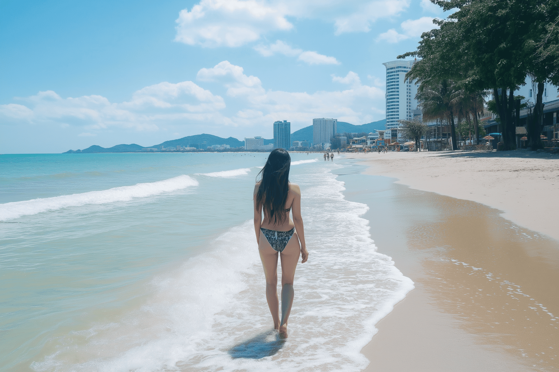A person with long hair strolls along a sunlit beach, where gentle waves kiss the shore. Tall buildings frame the background alongside distant mountains beneath partly cloudy skies, making this coastal area calm and picturesque.