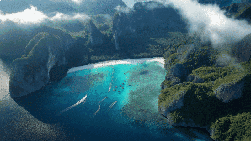 A stunning aerial view of the Phi Phi Islands reveals a tropical bay with turquoise waters, lush green cliffs, and boats leaving wake trails. The white sandy beach lines the bay, set against a backdrop of mountains and scattered clouds—inviting you to tour this paradise.