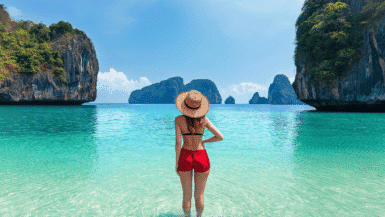 A woman in a sun hat and red shorts stands in shallow, clear turquoise water, savoring her tropical trip. She gazes at the large rocky cliffs and a distant island under the bright blue sky, cherishing this serene moment.