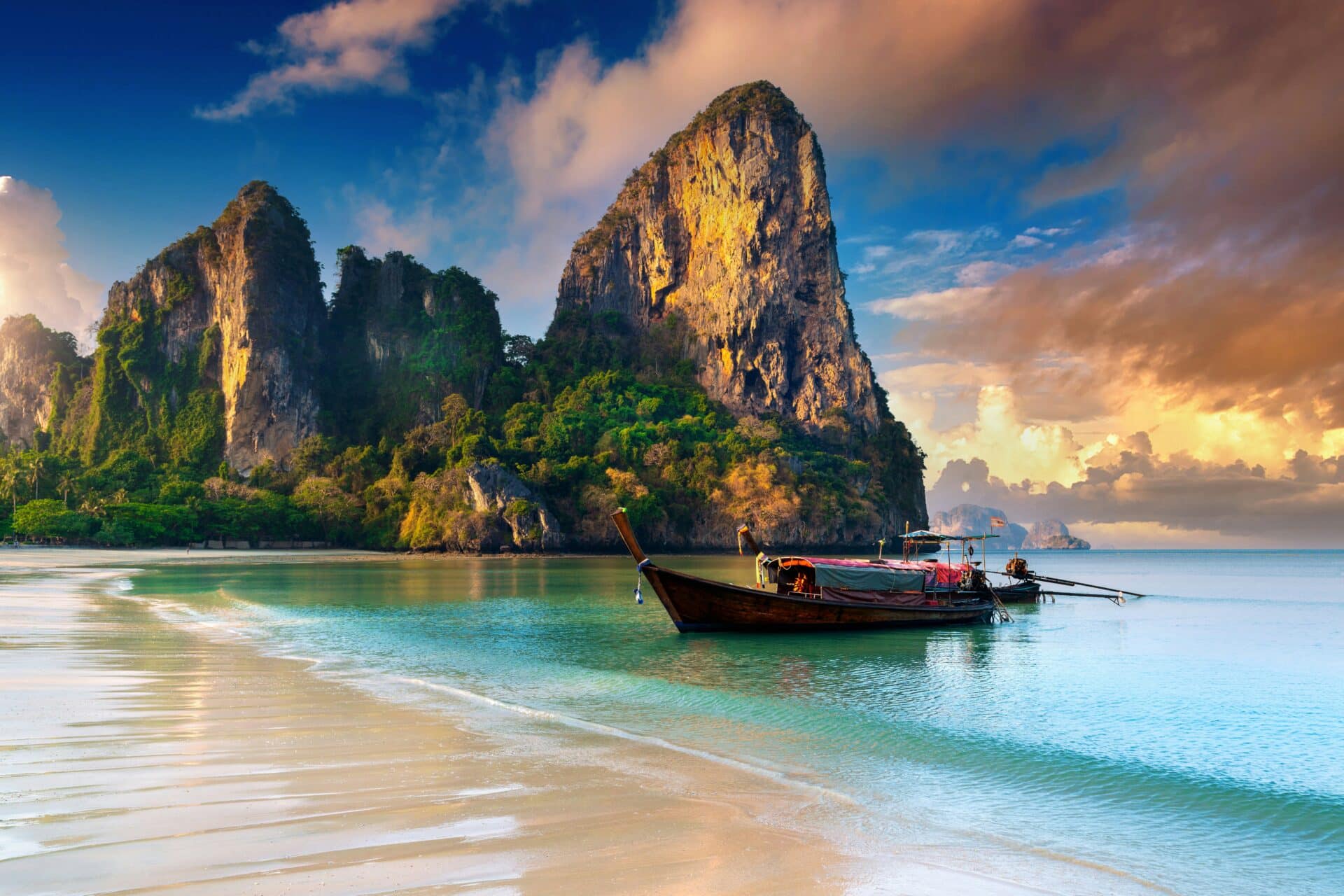 A traditional long-tail boat floats on the clear turquoise water near a sandy beach. Majestic limestone cliffs rise dramatically in the background under a partly cloudy sky, creating a serene and picturesque tropical scene, making it one of the Top 10 Things to Do in Krabi.