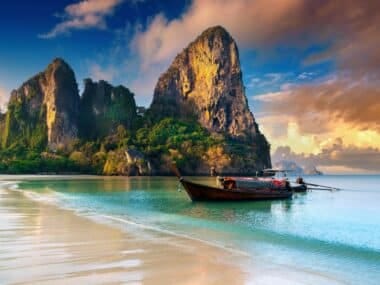 A traditional long-tail boat floats on the clear turquoise water near a sandy beach. Majestic limestone cliffs rise dramatically in the background under a partly cloudy sky, creating a serene and picturesque tropical scene, making it one of the Top 10 Things to Do in Krabi.