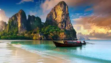 A traditional long-tail boat floats on the clear turquoise water near a sandy beach. Majestic limestone cliffs rise dramatically in the background under a partly cloudy sky, creating a serene and picturesque tropical scene, making it one of the Top 10 Things to Do in Krabi.