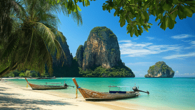 A serene beach scene with turquoise waters and towering limestone cliffs in the background beckons visitors during the best time to visit. Two wooden long-tail boats rest on the sand under lush tropical trees, offering a perfect escape beneath a clear blue sky.