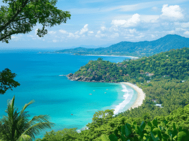 A scenic view of Phuket's tropical coastline with turquoise waters and a white sandy beach. Lush green hills surround the area, and a few boats dot the water. Under a bright sky scattered with fluffy clouds, its clear why many consider this paradise the best time to visit.