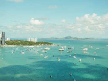 Aerial view of a coastal city with numerous boats scattered in the turquoise sea. The city features tall buildings along the shore, and distant green hills under a partly cloudy sky. Its easy to see why many say the best time to visit Pattaya is when this vibrant scene comes alive.