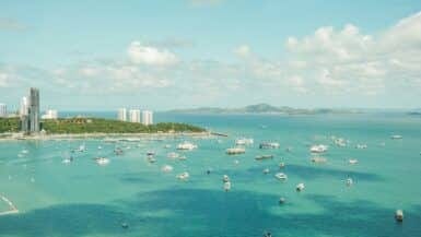 Aerial view of a coastal city with numerous boats scattered in the turquoise sea. The city features tall buildings along the shore, and distant green hills under a partly cloudy sky. Its easy to see why many say the best time to visit Pattaya is when this vibrant scene comes alive.